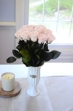 a white vase filled with pink roses sitting on top of a table next to a candle