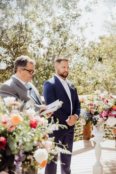 two men standing next to each other in front of flowers and greenery at a wedding ceremony