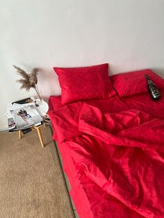 a bed with red sheets and pillows in a room next to a magazine on the floor