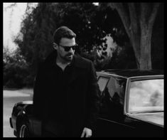 black and white photograph of a man walking next to a car in front of a tree