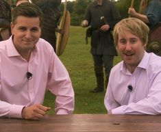 two men sitting at a wooden table in front of some people with shields on their heads