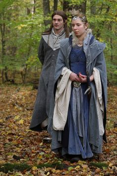 two people dressed in medieval clothing walking through the woods