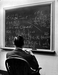 a man sitting in front of a blackboard with lots of calculations on it and writing on it