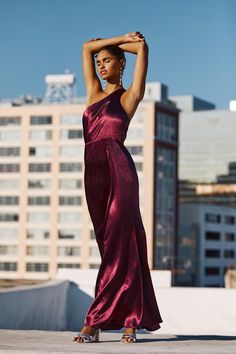 a woman in a long purple dress standing on top of a roof with her arms behind her head