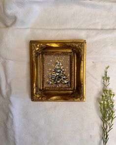 a small christmas tree in a gold frame on a white sheet with some green plants