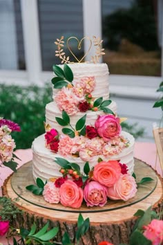 a wedding cake with pink flowers and greenery sits on a tree stump in front of a window