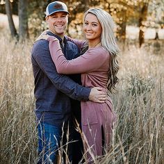 a man and woman standing in tall grass with their arms around each other, smiling at the camera