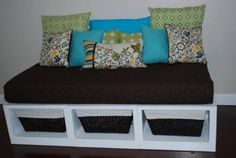 a day bed with baskets underneath it and pillows on the bottom shelf, in front of a white wall