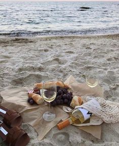 two glasses of wine, bread and grapes are on the sand at the beach by the water