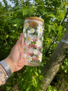 a hand holding a mason jar with daisies painted on the outside and inside, in front of some trees