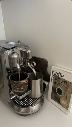 an espresso machine is on the counter next to a coffee cup and book