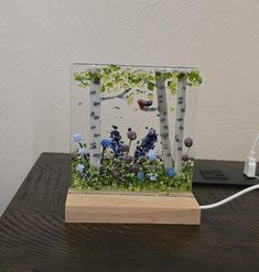 a glass block with flowers and trees in it on top of a wooden table next to a phone charger