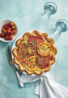 a pie with tomatoes and herbs on it next to some wine glasses, forks and spoons