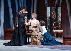 three women sitting at a table talking to each other in front of a tree filled room