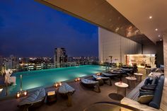 an outdoor swimming pool with lounge chairs and tables next to it at night in front of the cityscape