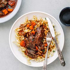 a white plate topped with meat and pasta next to a bowl of soup on a table