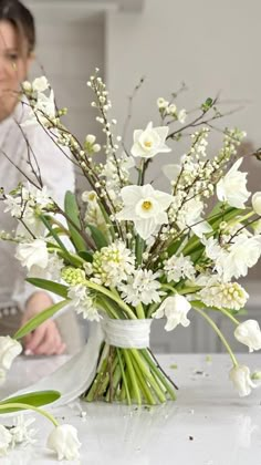 a woman arranging flowers in a vase on a counter top with other white flowers and greenery