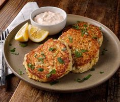 two crab cakes on a plate next to a bowl of sour cream and lemon wedges