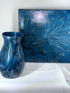 a blue vase sitting next to a painting on a white tablecloth with a white cloth underneath it