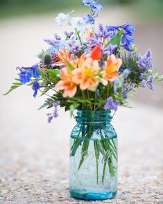 a blue mason jar filled with flowers on top of a gravel covered ground next to trees