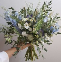 a person holding a bouquet of blue and white flowers with greenery in their hand