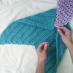 someone is knitting a blue knitted blanket on top of a white bed with a pink and green crochet design