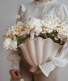 a woman holding a vase with white flowers in it's hands and wearing beige pants