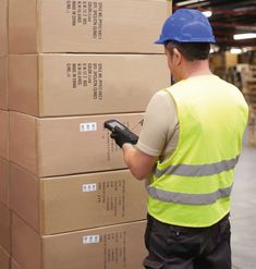 a man wearing a hard hat and safety vest is using a handheld device to check boxes in a warehouse