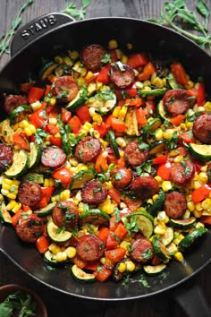 a skillet filled with sausage, corn and vegetables on top of a wooden table