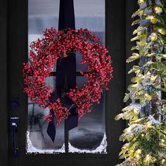 a red berry wreath on the front door of a house with christmas trees in the background