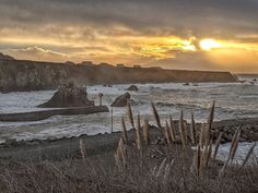 the sun is setting over the ocean with rocks in the foreground and grass on the shore