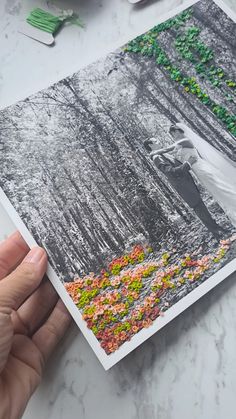 a person holding up a photo with trees and flowers in the background on a table