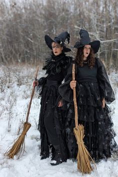 two women dressed as witches standing in the snow with brooms and wearing black clothes