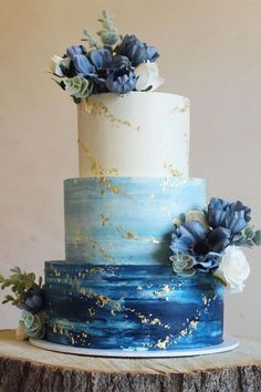 a three tiered blue and white cake with flowers on top is sitting on a tree stump