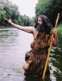 a man dressed in native garb holding a stick and standing in the water with his arms outstretched