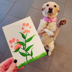 a dog is standing on its hind legs while holding up a piece of paper with flowers painted on it