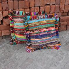 two colorful pillows sitting next to each other on the ground in front of a pile of bricks