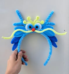 a hand is holding up a blue and yellow knitted headband with an orange nose