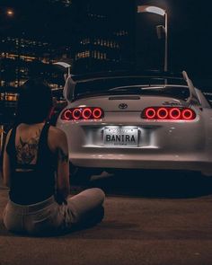 a woman sitting on the ground next to a white sports car in front of a city at night