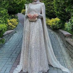 a woman in a silver gown standing on a brick walkway with trees and bushes behind her