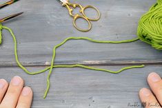 two hands holding yarn and scissors on a table next to green crochet needles