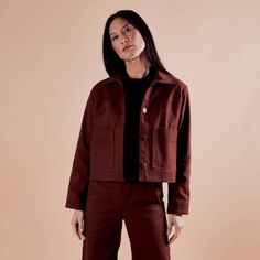 a woman standing in front of a pink wall wearing a brown jacket and black top