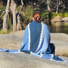 a woman wrapped in a blue blanket sitting on top of a rock next to a river