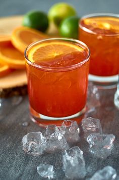 two glasses filled with liquid and ice sitting on top of a table next to sliced oranges