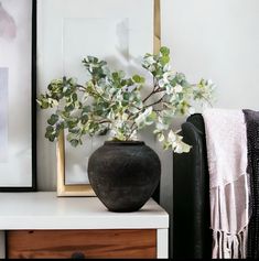 a black vase with white flowers sitting on a table next to a framed photograph and a blanket