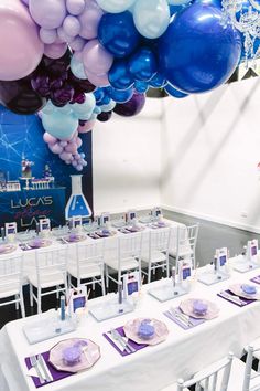 there are many balloons and plates on the table at this party with purple, blue, and white decorations