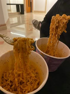 two bowls filled with noodles being held up by someone's hand, while another person sits on the floor in the background