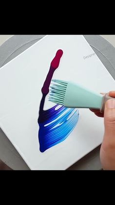 someone using a brush to paint the design on a square piece of paper with blue and red acrylic