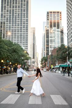 a man and woman holding hands crossing the street in front of tall buildings on a sunny day