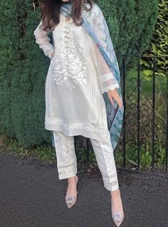 a woman standing in front of a fence wearing a white dress and blue shawl